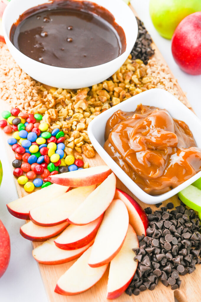 A caramel apple board. A wooden serving board with chocolate and caramel sauces in bowls, scattered with sliced apples and other apple toppings like nuts, mini chocolate chips, and mini M&Ms.