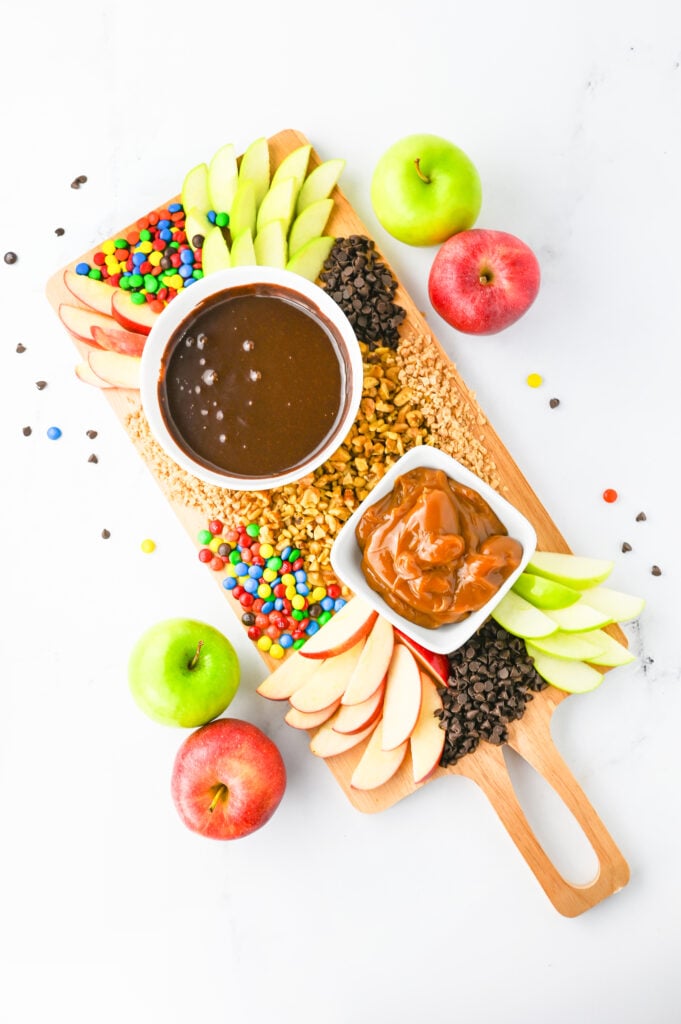 A caramel apple board. A wooden serving board with chocolate and caramel sauces in bowls, scattered with sliced apples and other apple toppings like nuts, mini chocolate chips, and mini M&Ms.