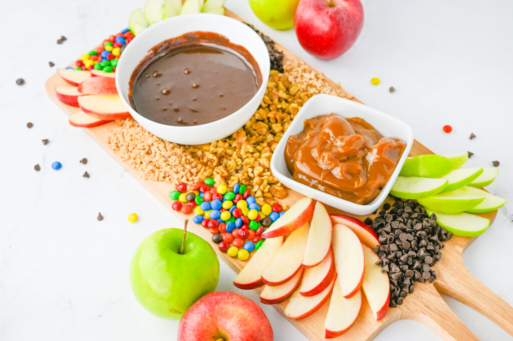 A caramel apple board. A wooden serving board with chocolate and caramel sauces in bowls, scattered with sliced apples and other apple toppings like nuts, mini chocolate chips, and mini M&Ms.