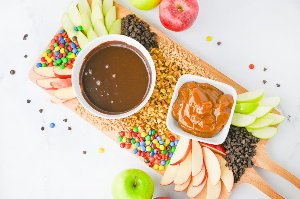 A caramel apple board. A wooden serving board with chocolate and caramel sauces in bowls, scattered with sliced apples and other apple toppings like nuts, mini chocolate chips, and mini M&Ms.