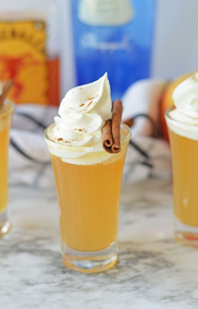 Apple pie shot in a clear glass, topped with a swirl of whipped cream and a cinnamon stick, set on a marble surface with blurred bottles and fall-themed decor in the background.