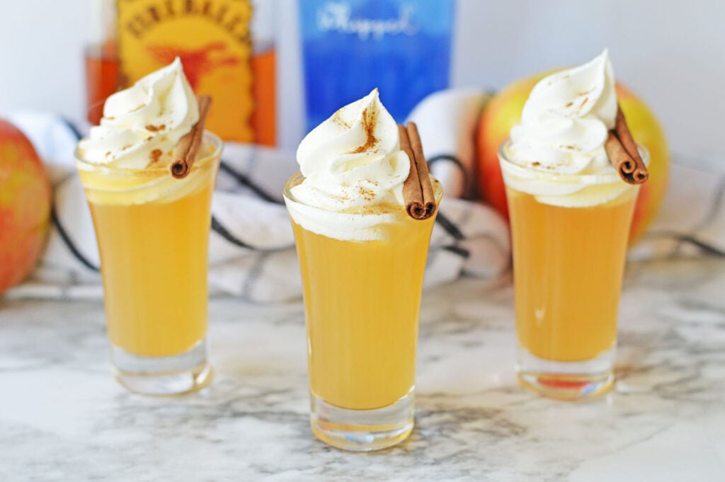 Three tall apple pie shots, topped with whipped cream and cinnamon sticks, arranged on a light countertop with fresh apples and bottles behind.