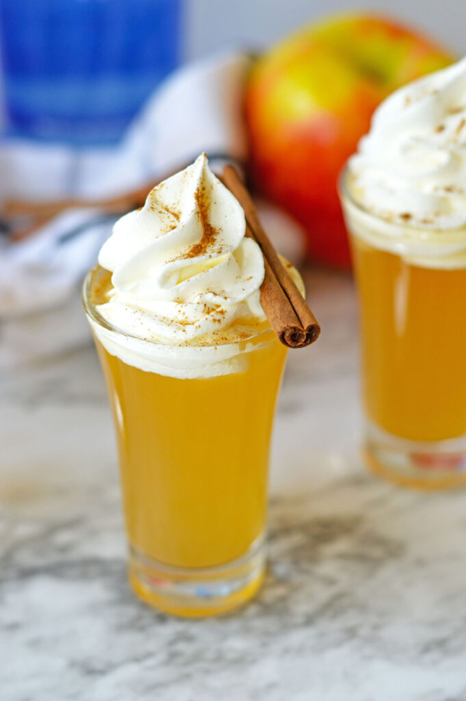 Close‑up of a single apple pie shot glass topped with a swirl of whipped cream, sprinkled cinnamon, and a cinnamon stick garnish.