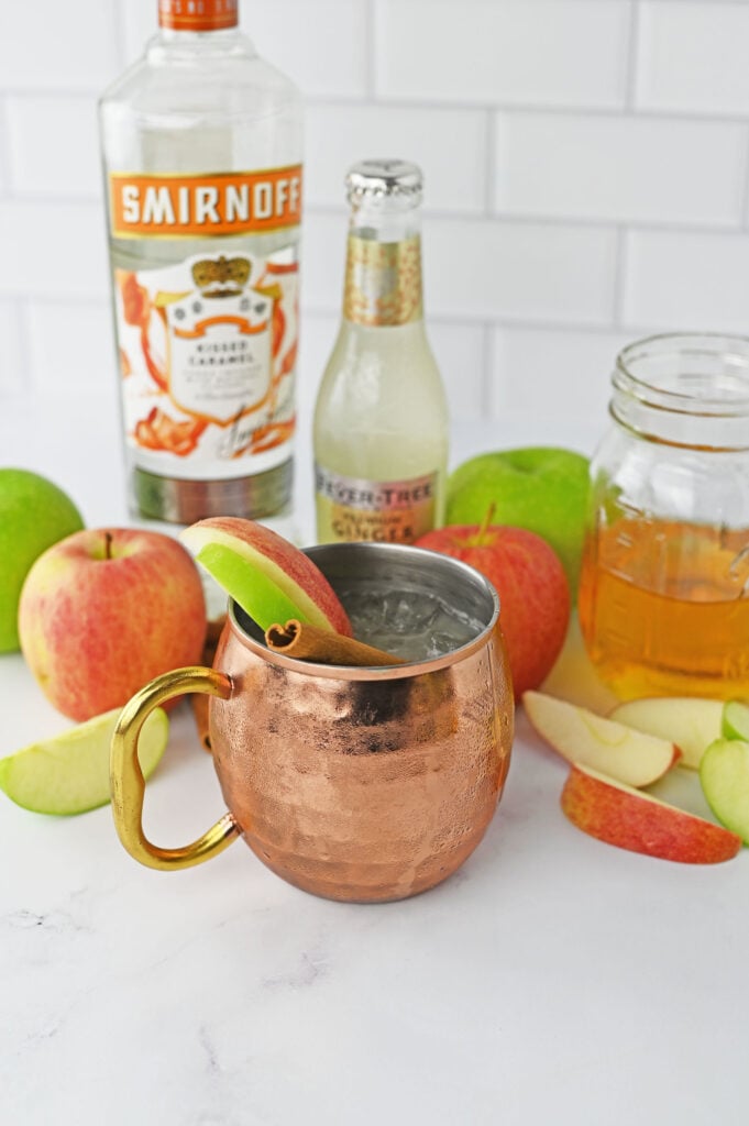 A caramel apple mule in a copper cup garnished with green and red apple slices and a cinnamon stick.