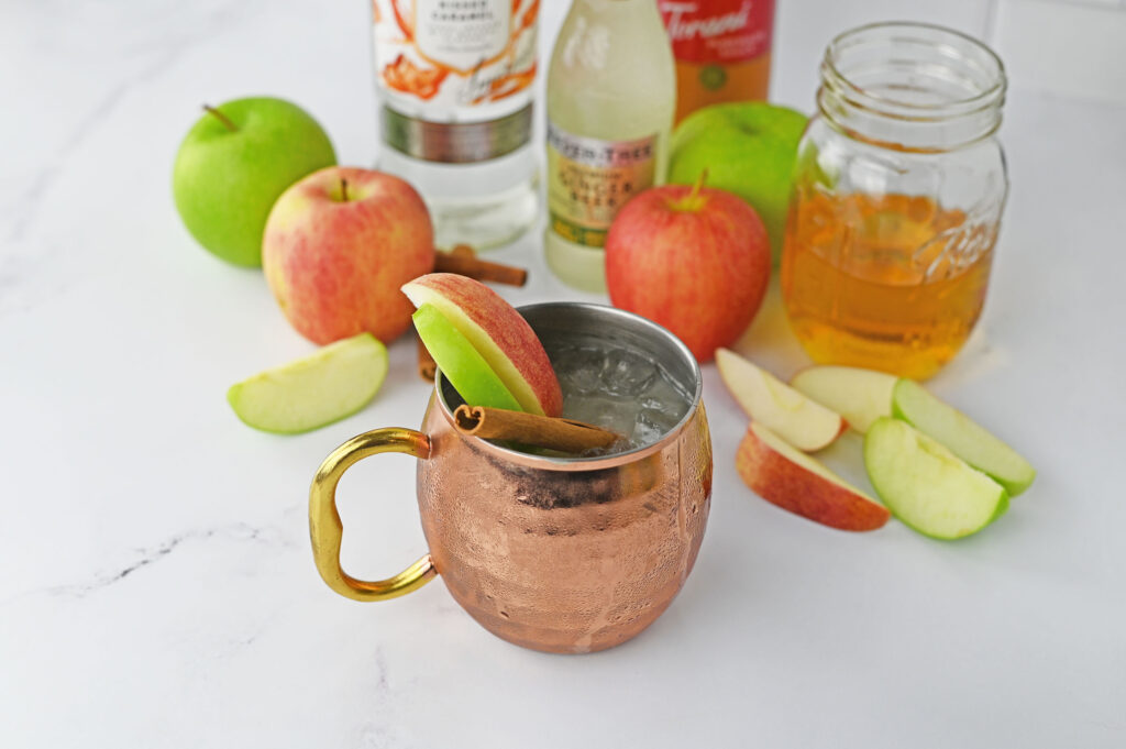 A caramel apple mule in a copper cup garnished with green and red apple slices and a cinnamon stick.