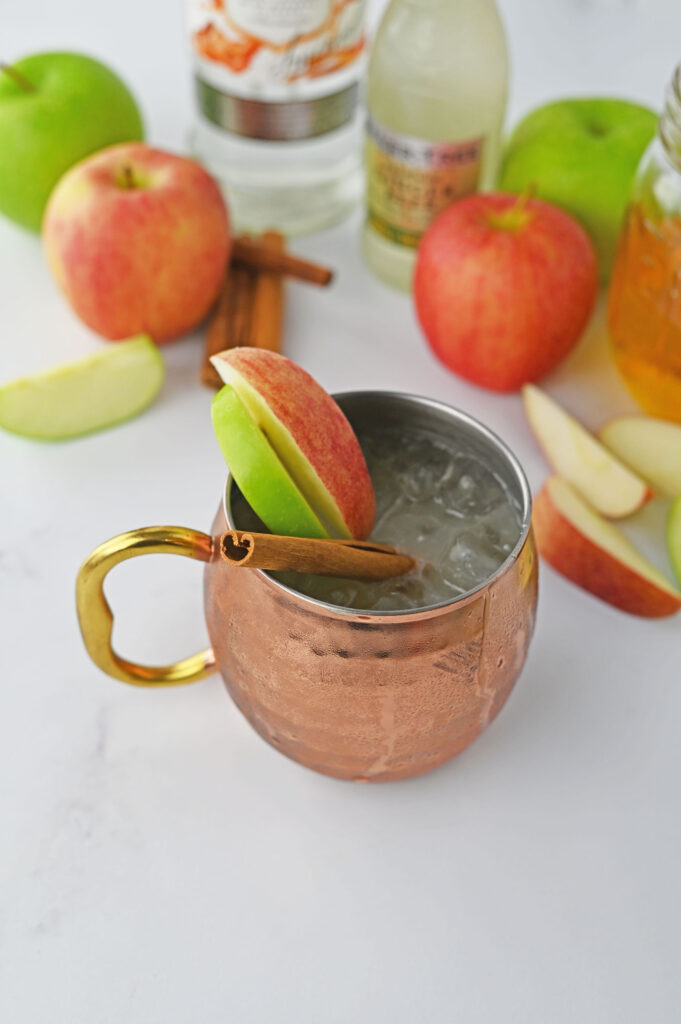 A caramel apple mule in a copper cup garnished with green and red apple slices and a cinnamon stick.