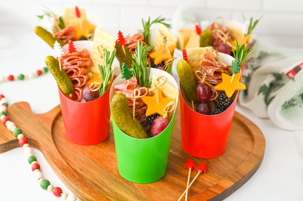 Festive red and green paper cups filled with charcuterie ingredients on a wooden board.