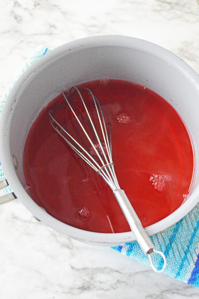 Red jello in a saucepan with a whisk.