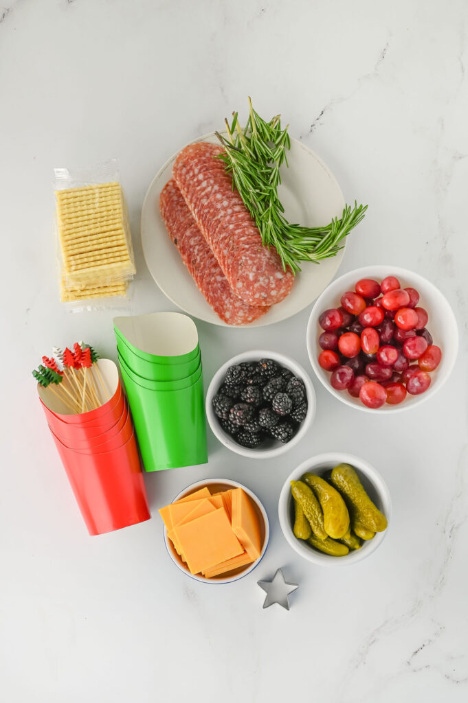 Christmas charcuterie cup ingredients- club crackers, salami, rosemary sprigs, blackberries, grapes, cheese, and gherkins with a stack of red and stack of green cups, and a star cookie cutter.