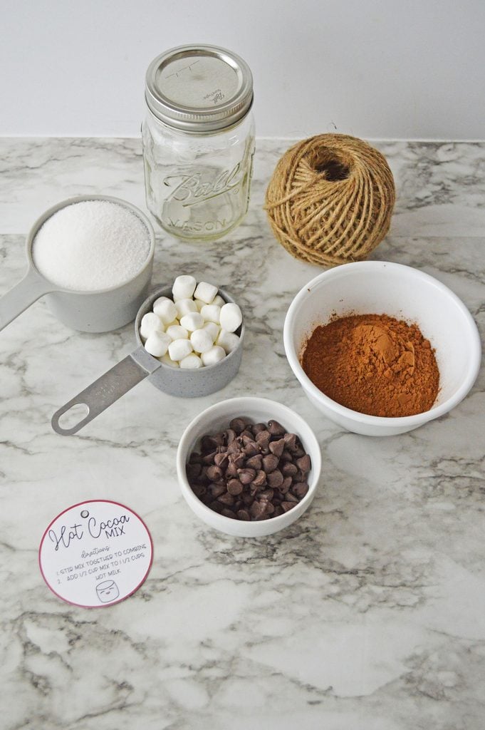 Ingredients for homemade hot cocoa in a jar including cocoa powder, sugar, chocolate chips, and marshmallows
