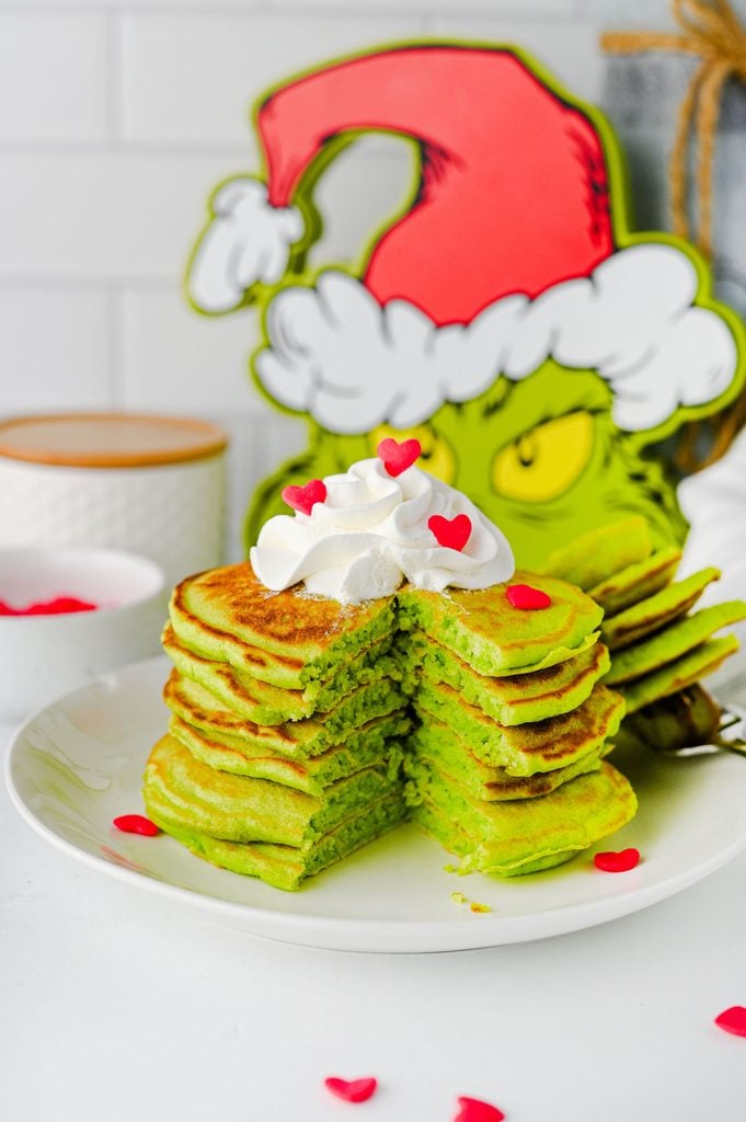 Stack of fluffy green Grinch pancakes topped with whipped cream and a red heart sprinkle.
