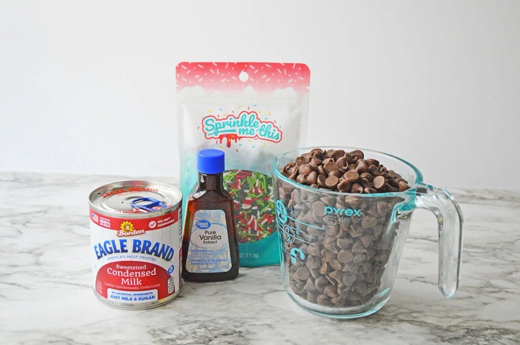 Ingredients for homemade Christmas fudge including chocolate chips, sweetened condensed milk, vanilla, and sprinkles.