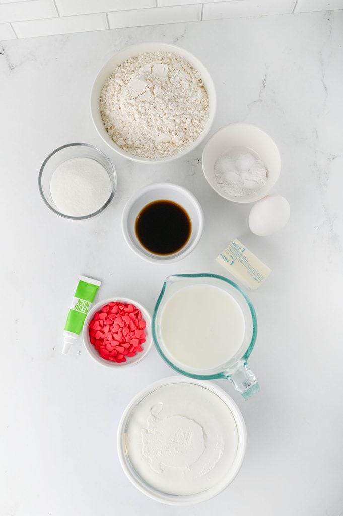 Ingredients for homemade Grinch pancakes including flour, sugar, baking powder, baking soda, milk, butter, egg, vanilla, green food coloring, and festive toppings.