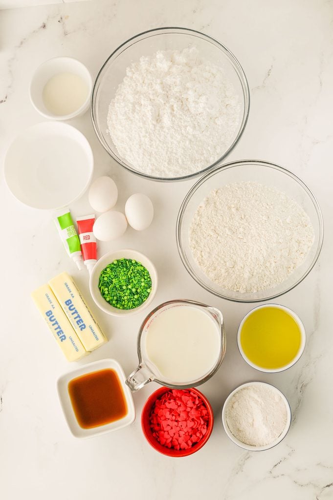 Ingredients for Grinch Poke Cake, including white cake mix, cheesecake instant pudding, butter, powdered sugar, milk, vanilla extract, green and red food coloring, and festive Christmas sprinkles.