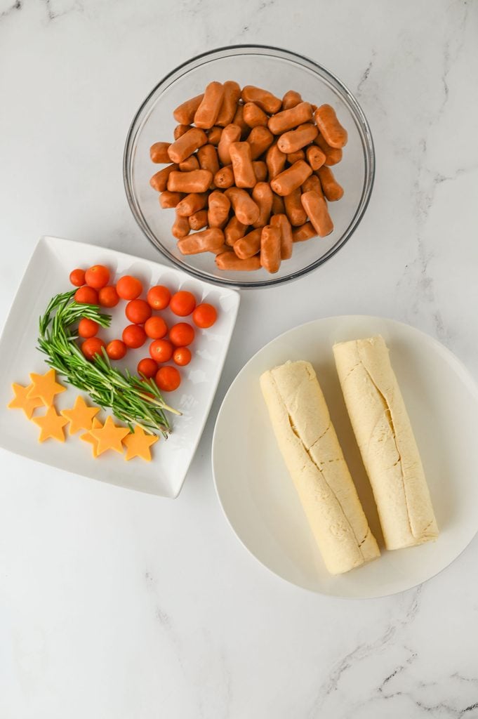 Ingredients for Christmas pigs in a blanket wreath, including crescent rolls, little smokies, fresh rosemary, cheddar cheese slices, and cherry tomatoes.