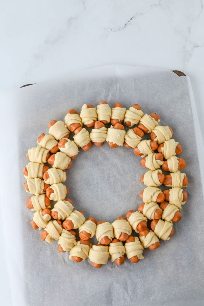 Unbaked pigs in a blanket arranged in a circular wreath shape on a parchment-lined baking sheet.